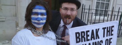 Two protestors, one in a bowler hat and smoking a cigar, another with the flag of Greece painted on their face and with chains wrapped around their neck. They hold a banner which reads 'break the chains of debt'.