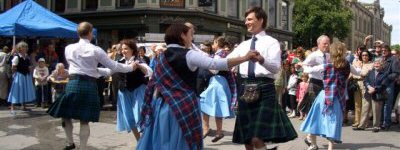 A group of people in traditional Scottish dress, including kilts and sashes, doing country dancing in the street.